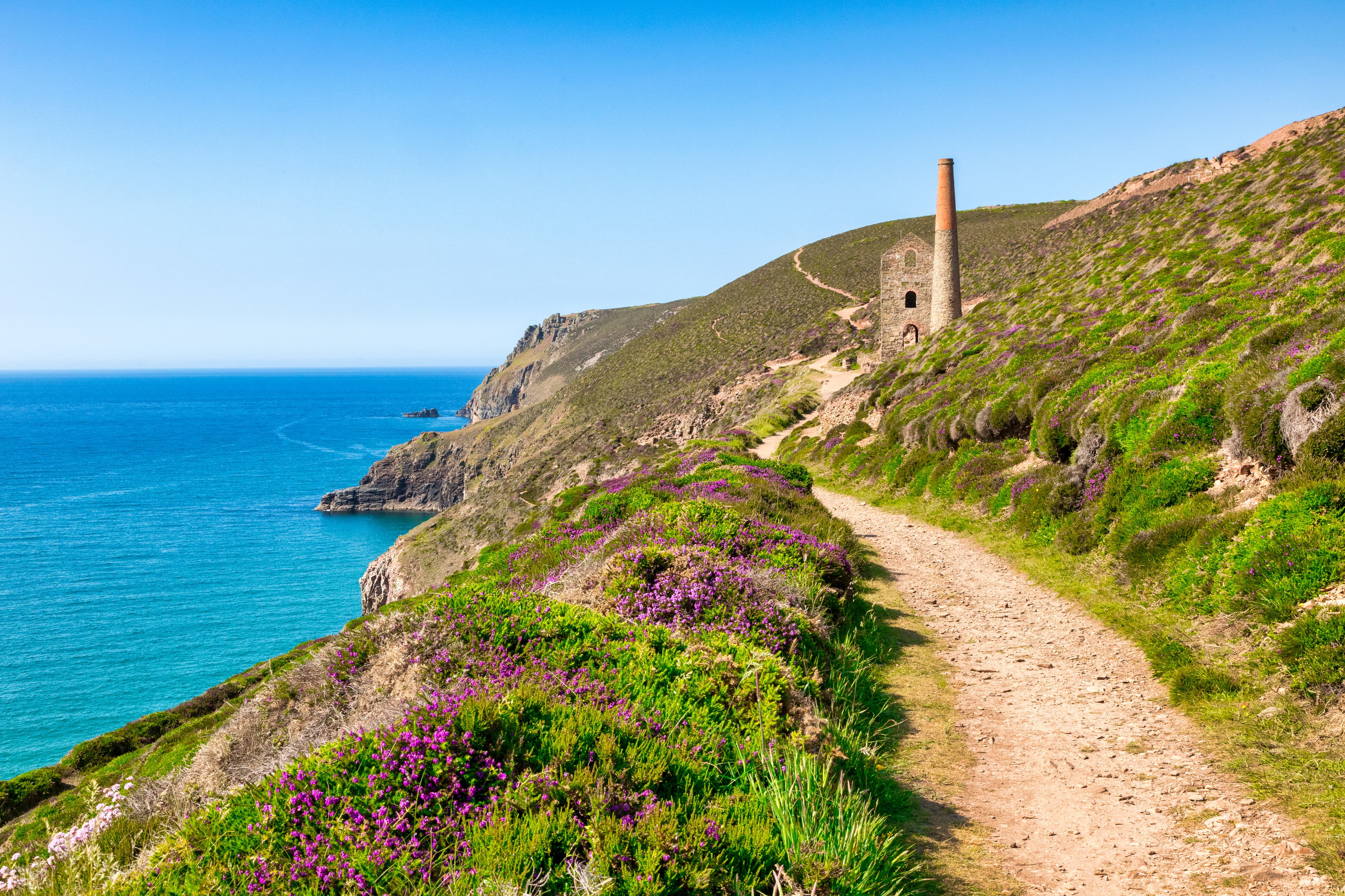 A coastal pathway with greenery surrounding the path, ocean on the left, on the right a hill rises, on the path a brick structure with a chimney can be seen