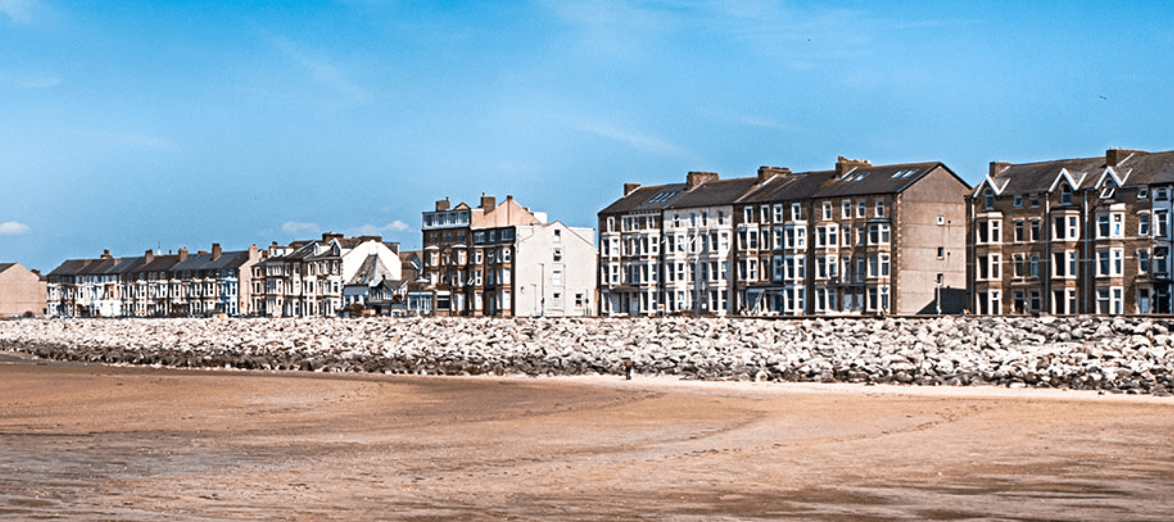 Row of coastal buildings behind a rock barrier, with a sandy beach in the foreground and a clear blue sky above.