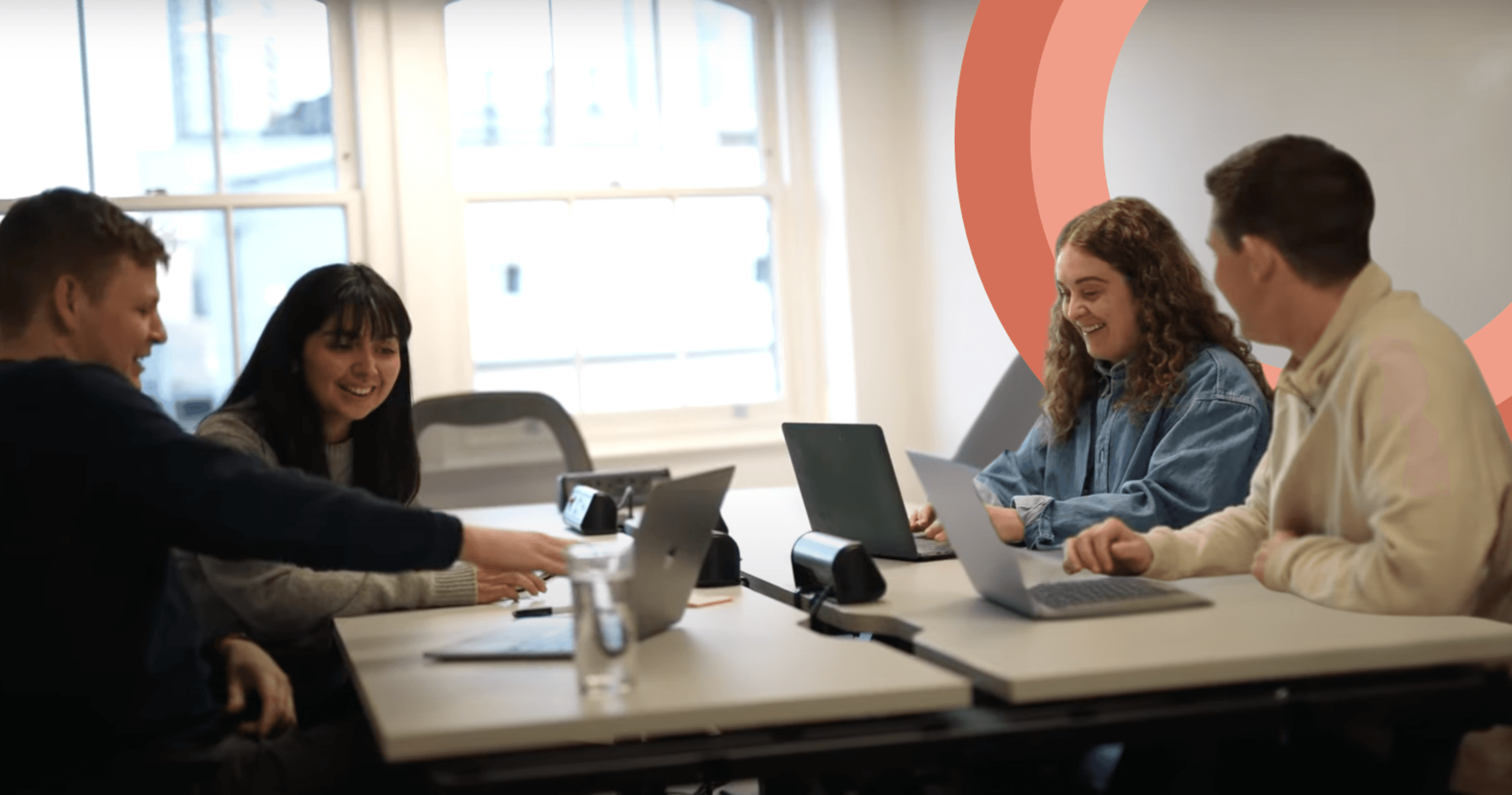 Four people sitting at a table with laptops, smiling and talking in a bright office space.