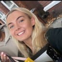 A smiling person with long blonde hair holds shopping bags outdoors near a brick building.